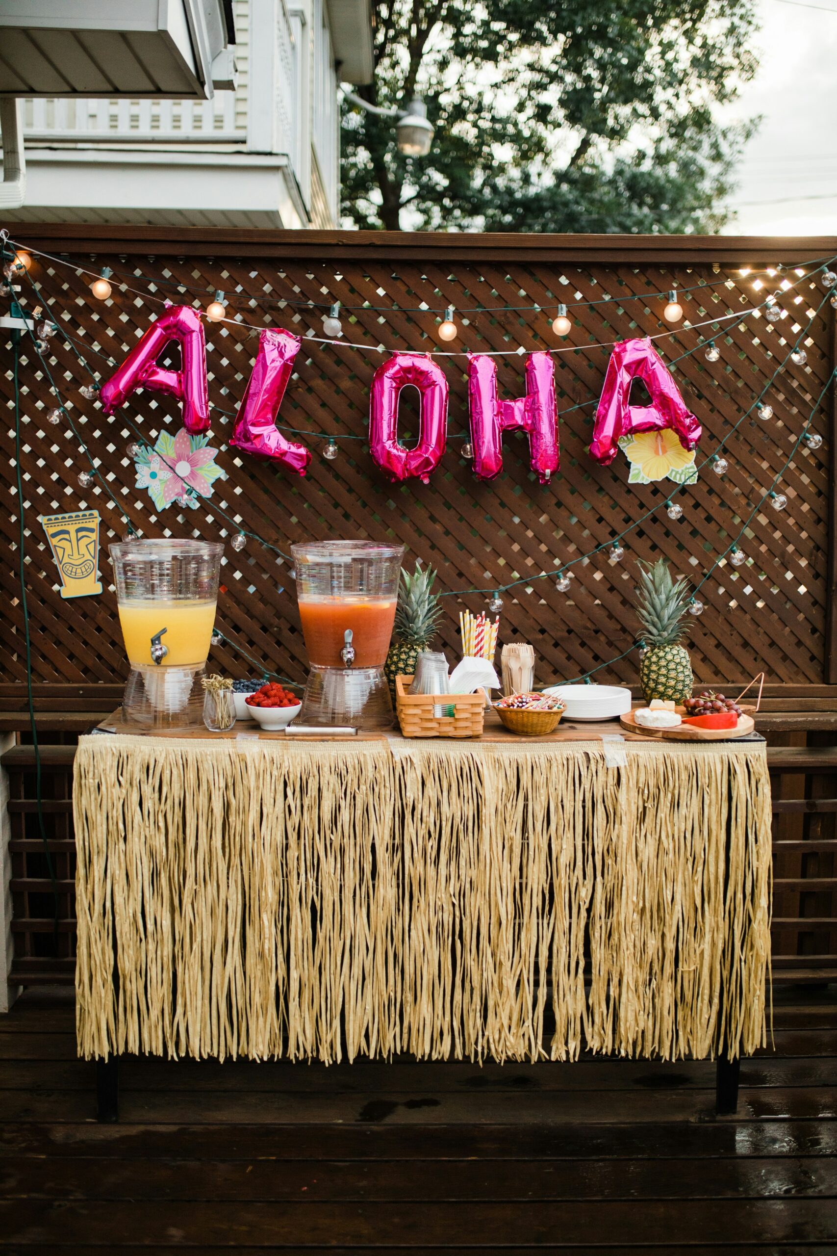 Outdoor luau party table with tropical drinks, pineapples, and pink aloha balloons.