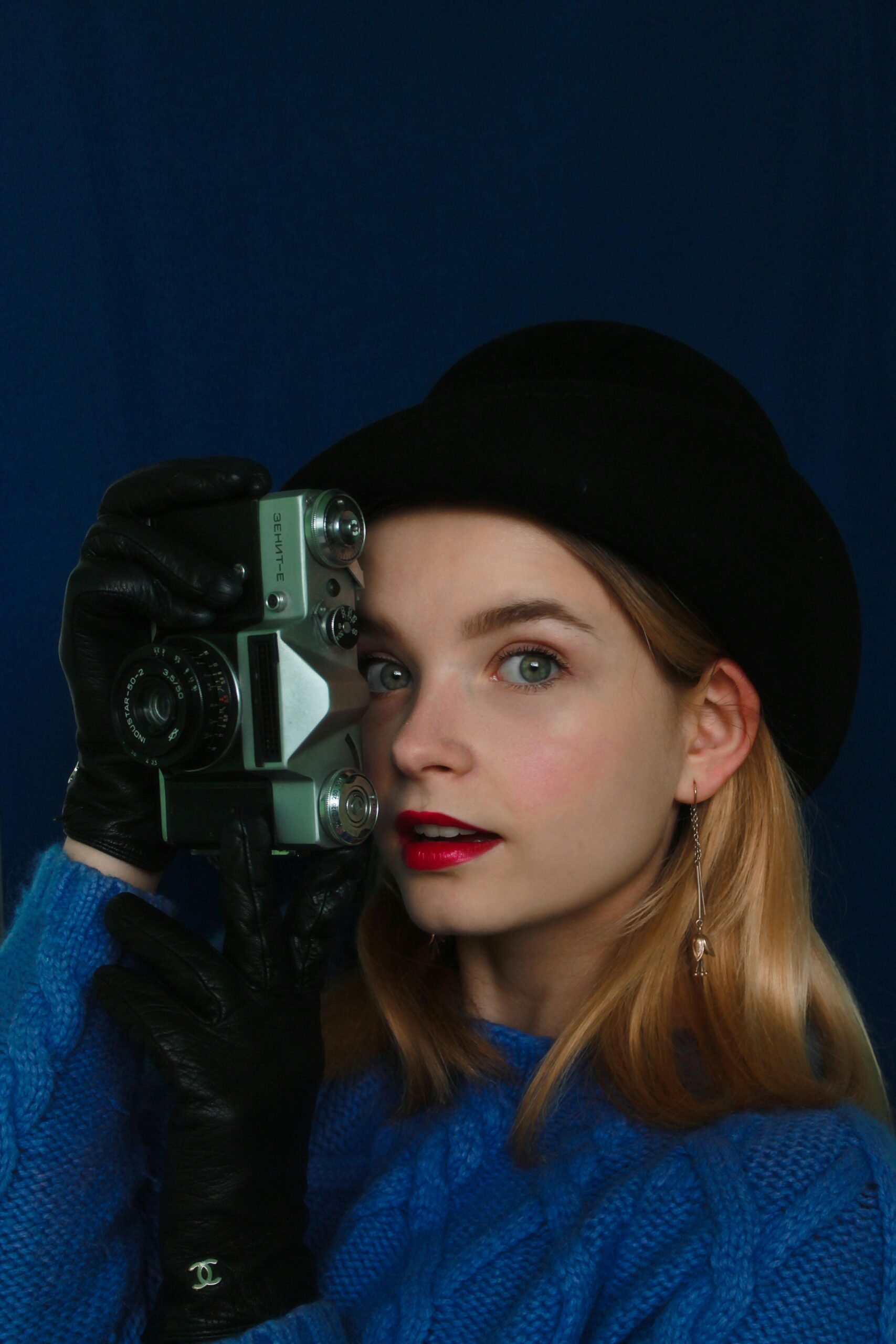 Girl holding a vintage camera while wearing a black hat and gloves against a dark background.