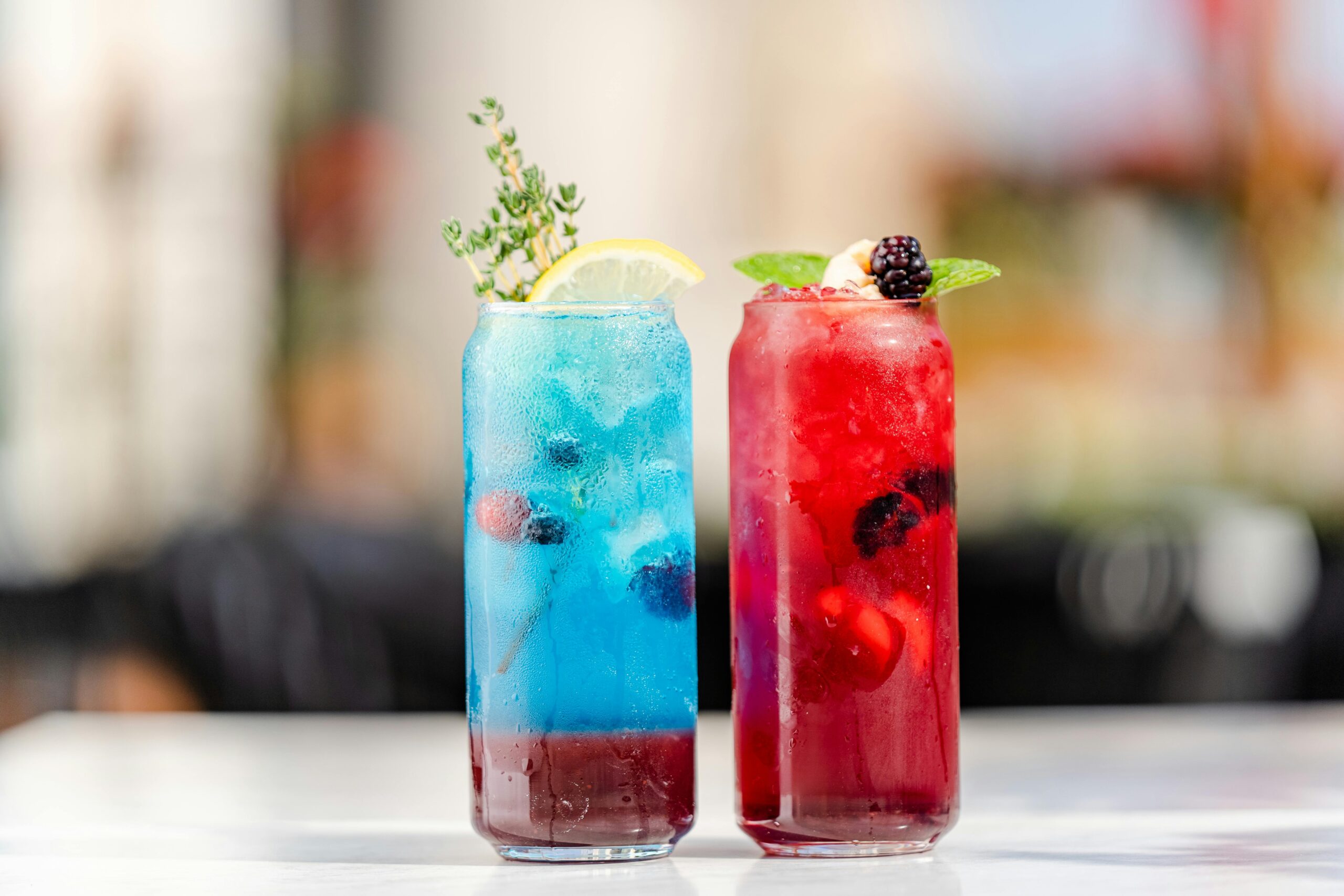 Two colorful drinks in glasses garnished with fruit and herbs on a table.