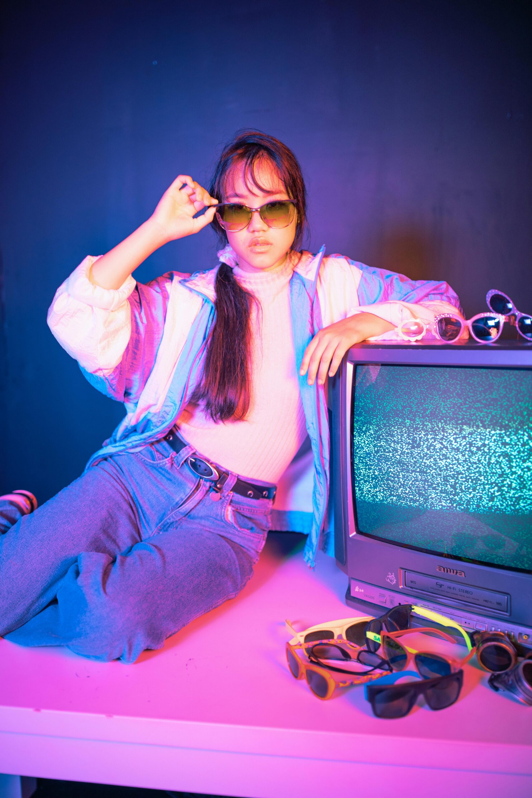 Teen girl in retro outfit posing with sunglasses beside a vintage TV and neon lighting.
