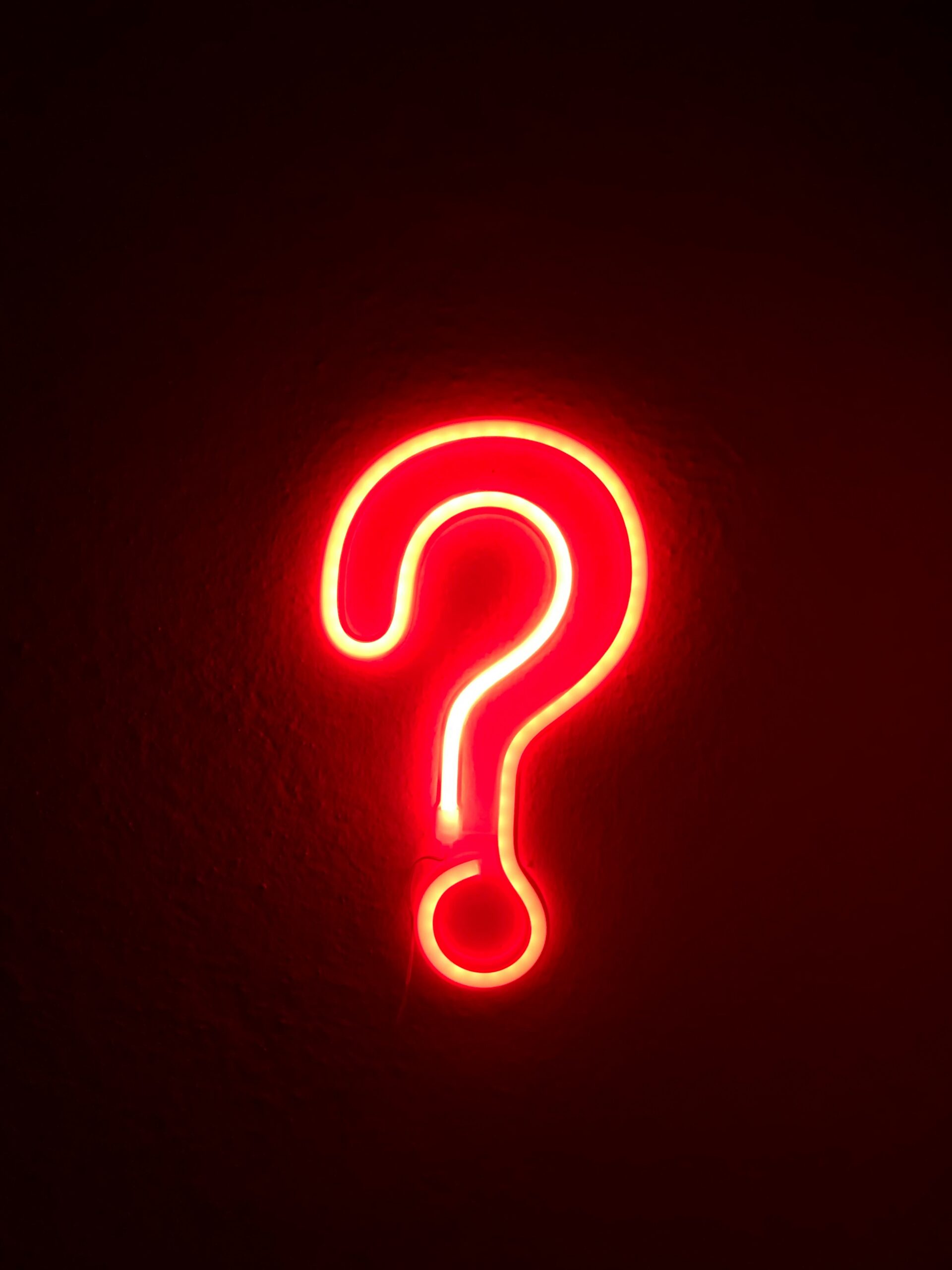 Glowing red neon question mark sign on a dark wall.