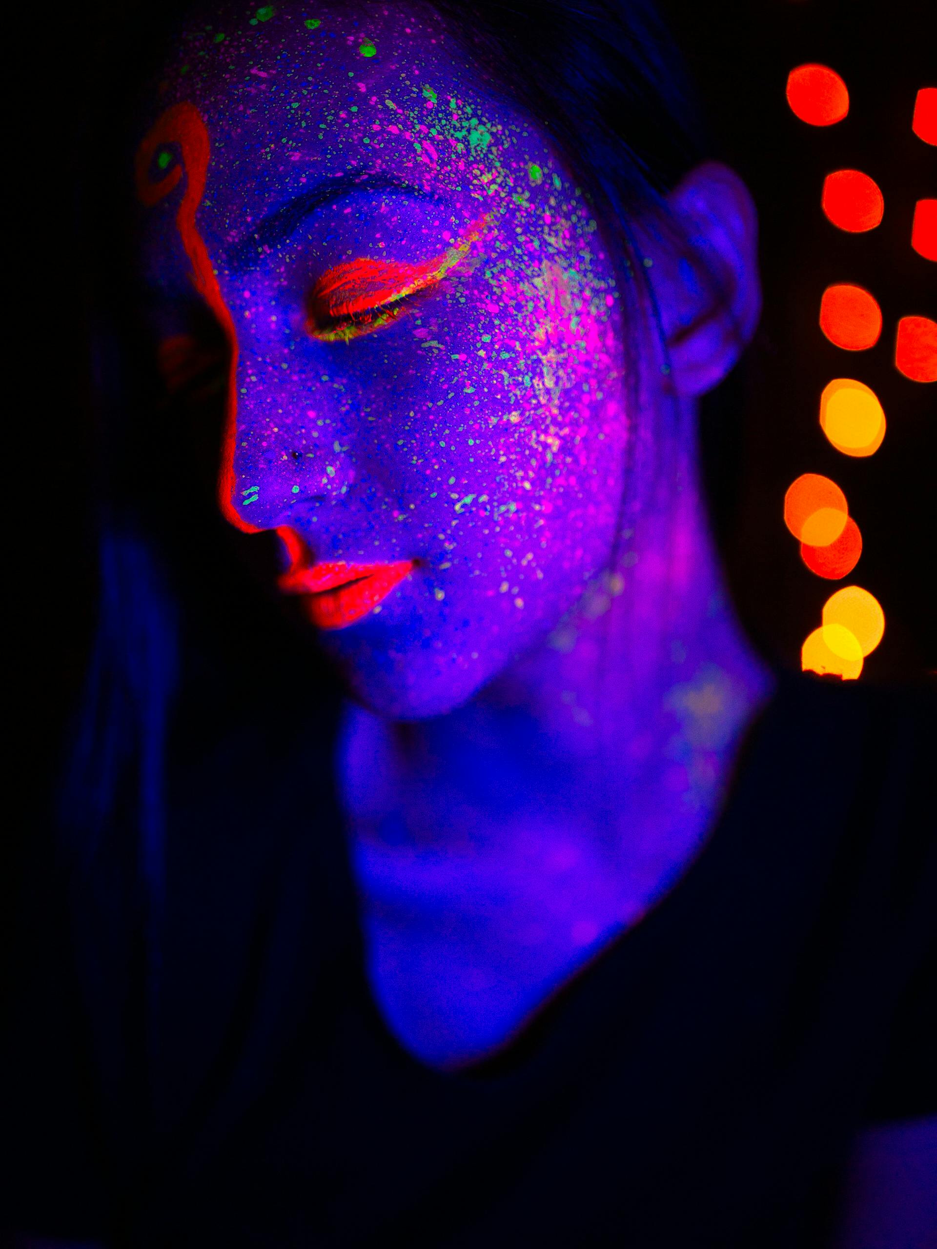 Person with glowing neon face paint under blacklight with colorful lights in the background.