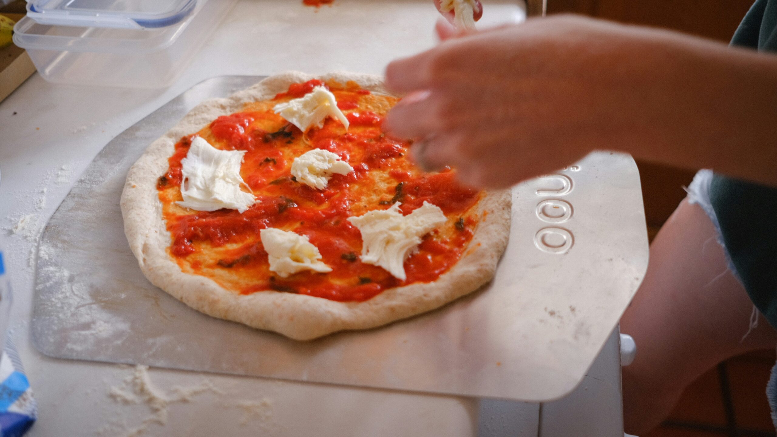 Hands adding cheese to homemade pizza dough with tomato sauce on a kitchen counter.