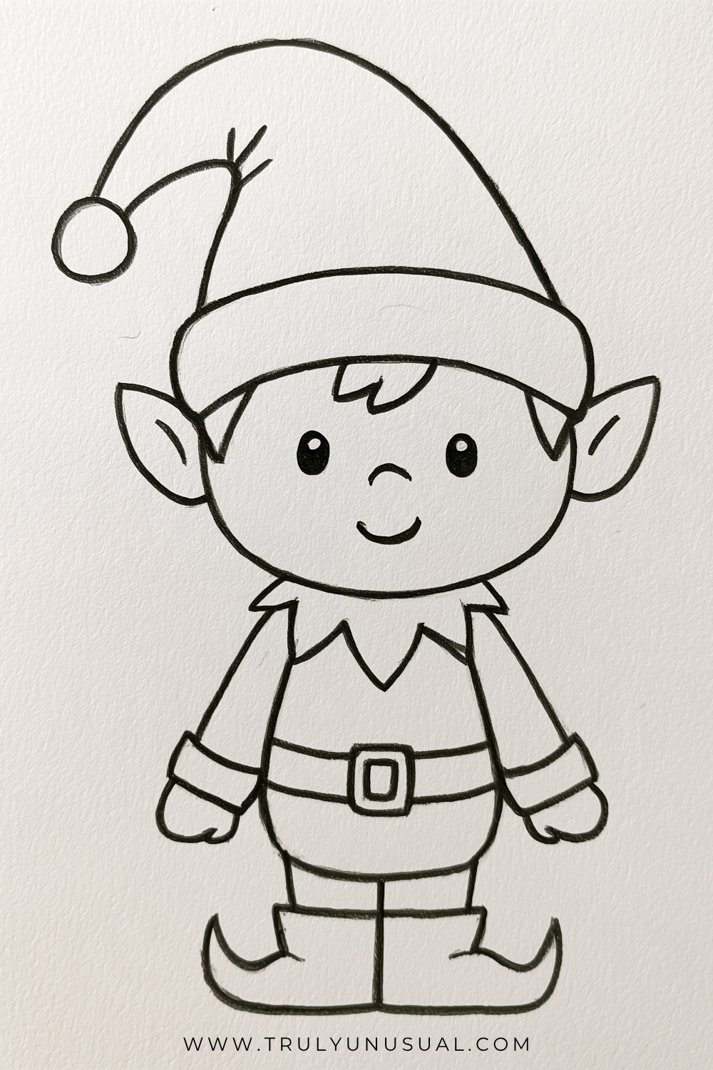 Cute and easy Christmas elf drawing featuring a smiling little elf with a large round face, pointed ears, big oval eyes, and a small tuft of hair peeking out from beneath an oversized Santa-style hat with a round pom-pom. The elf wears a festive outfit with a pointed collar, long sleeves, a wide belt with a square buckle, and oversized shoes with curled, pointed toes. Simple black-and-white hand-drawn illustration with bold pencil outlines on textured white paper, suitable for beginner Christmas drawing ideas.