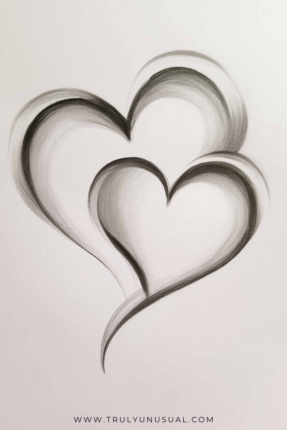 Monochrome drawing of two overlapping hearts with broad, softly shaded ribbon-like edges. A larger heart rises behind a smaller heart at the lower right; dark inner contours fade into pale centres, and a long curved point sweeps downwards beneath them.